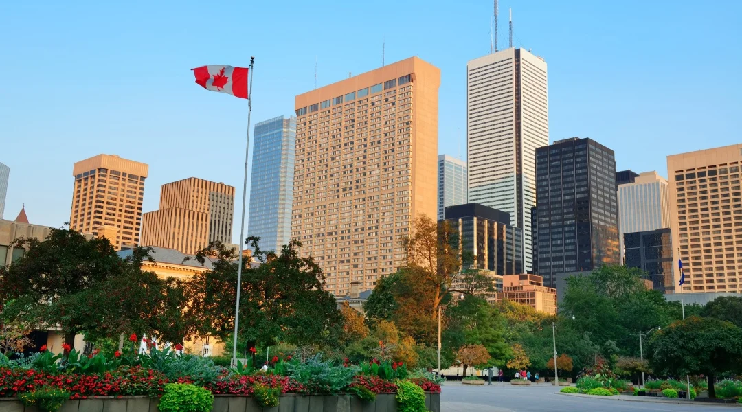 City skyline featuring tall buildings with a prominent Canadian flag waving in the foreground.